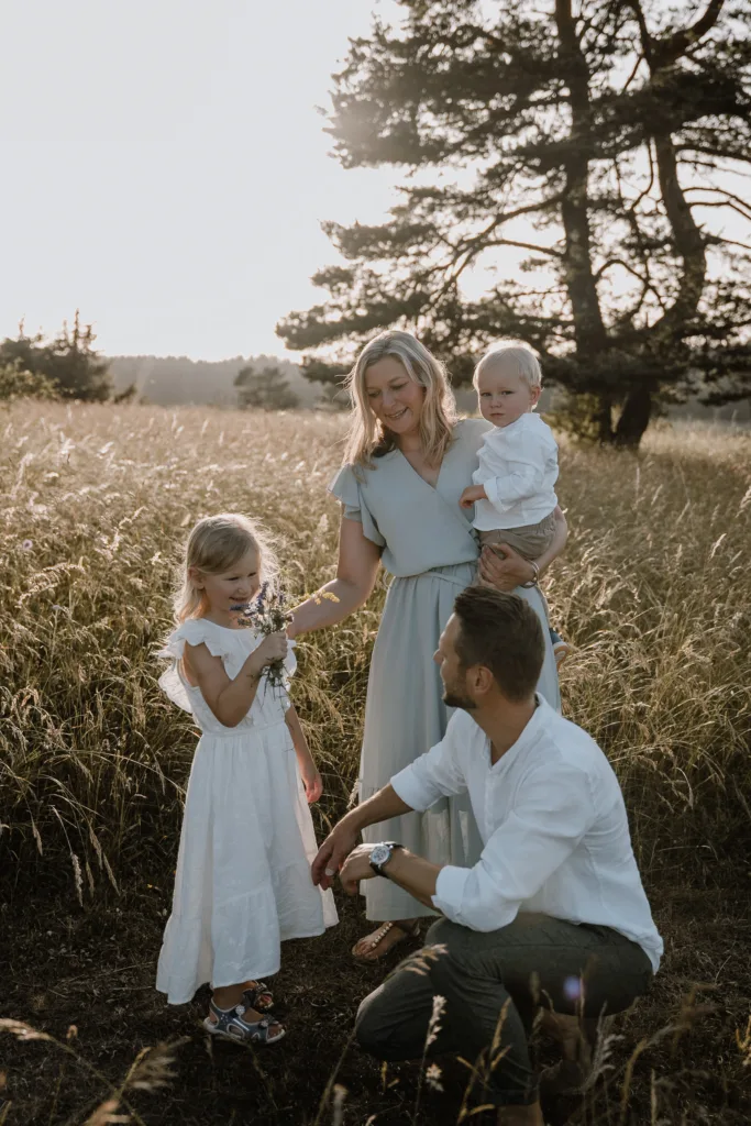 Familie mit zwei Kindern auf einer sommerlichen Wiese, Eltern knien vor ihrer Tochter mit Blumenstrauß.