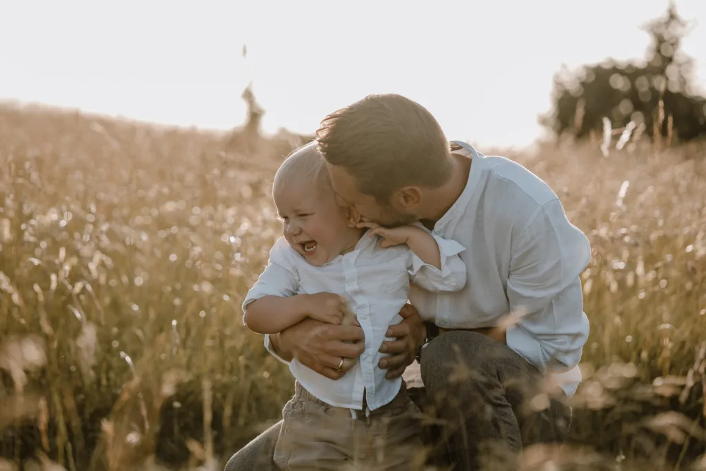 Vater hält lachenden Sohn im hohen Gras und küsst ihn liebevoll.