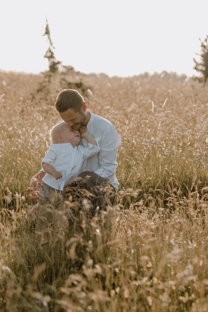 Vater sitzt mit Kleinkind im hohen Sommergras und beide lachen.