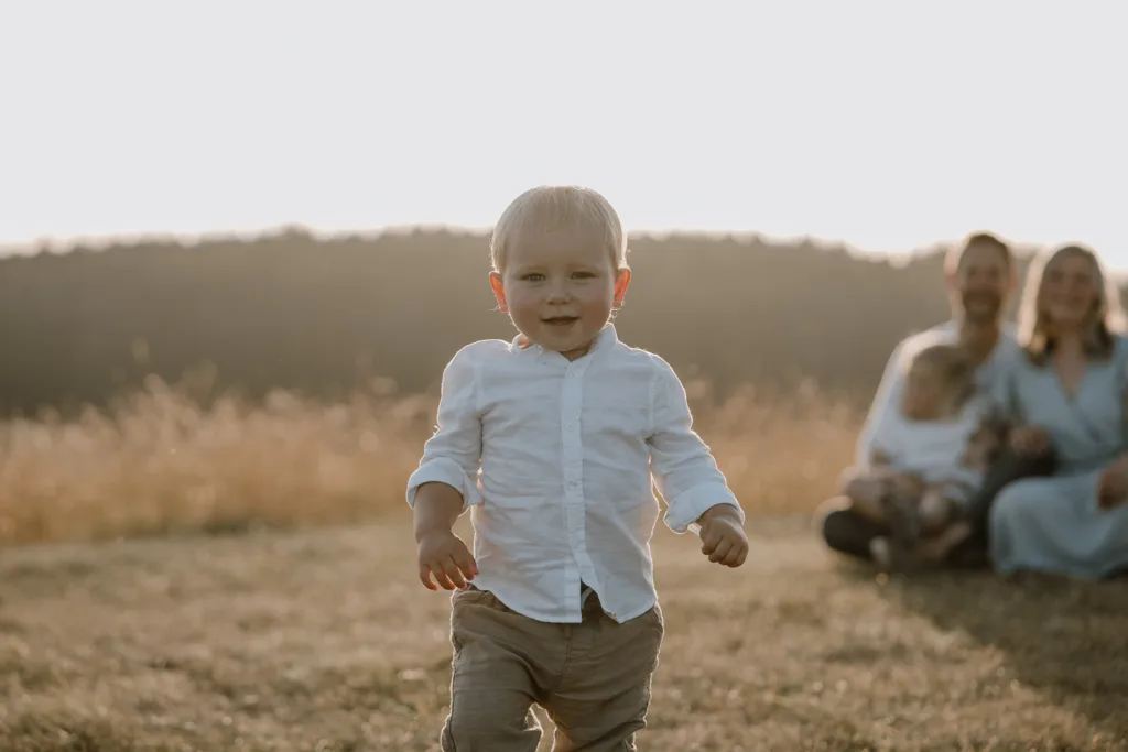 Kleinkind läuft lachend auf der Wiese auf die Kamera zu, Eltern im Hintergrund.