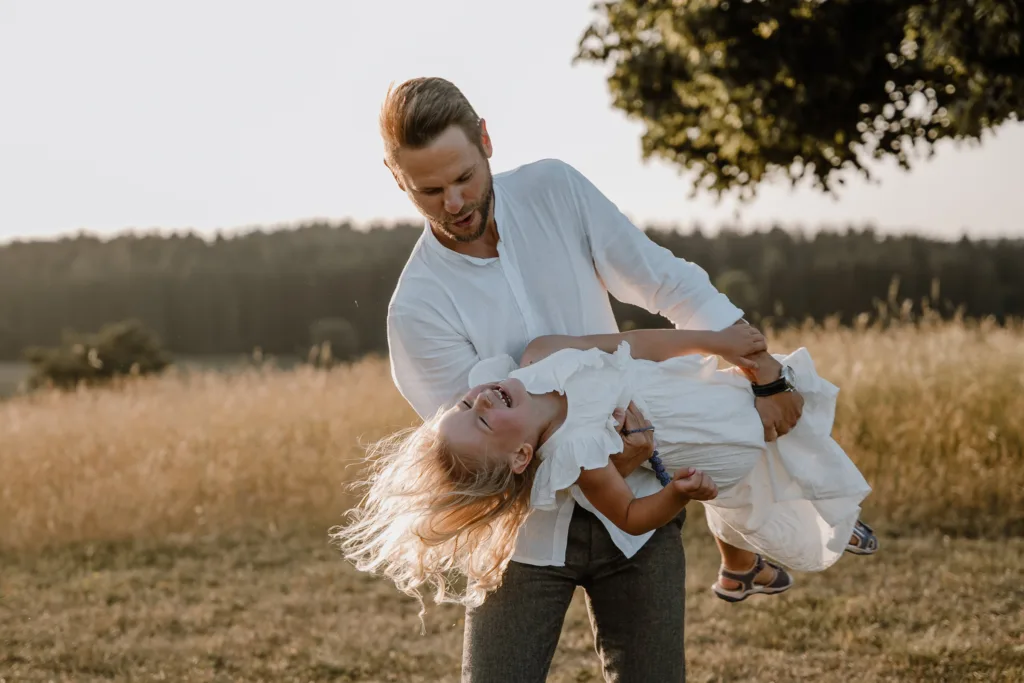 Vater hält Tochter spielerisch im Arm, sie lacht ausgelassen im Abendlicht.