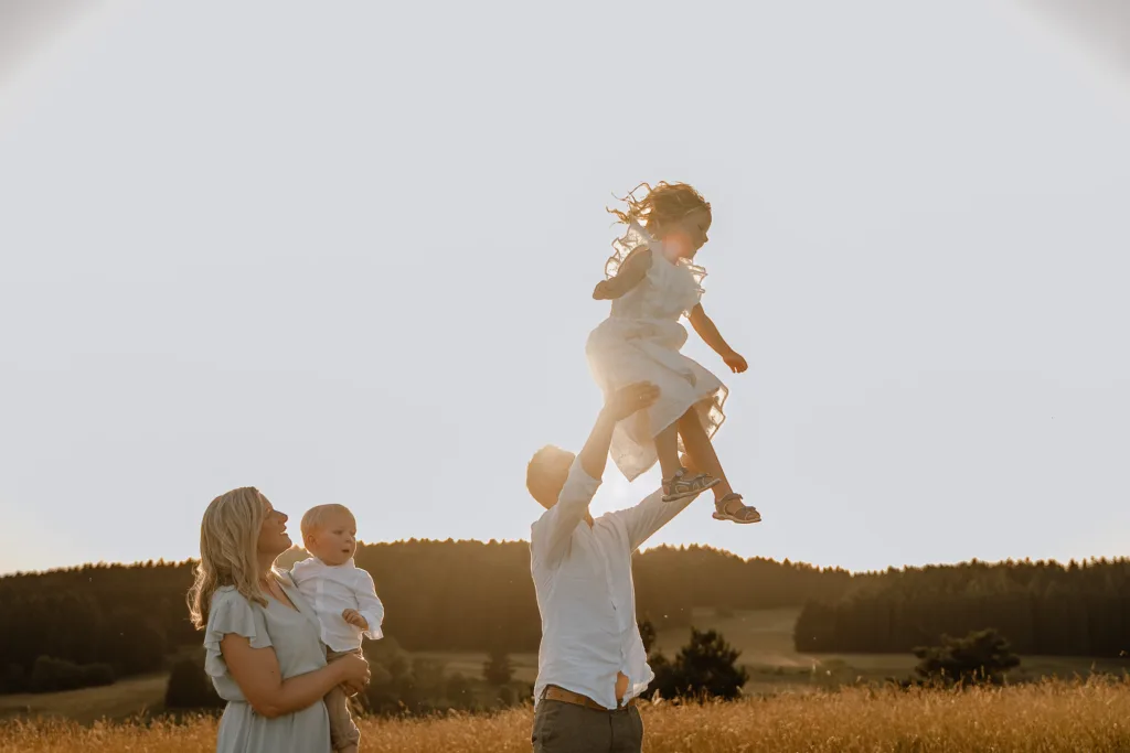 Vater hebt Tochter hoch, Mutter steht daneben mit Baby in der Natur.