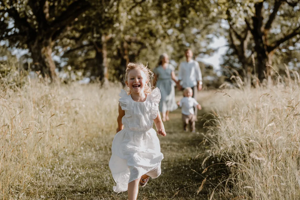 Lachendes Mädchen läuft barfuß über einen Wiesenweg, Familie unscharf im Hintergrund