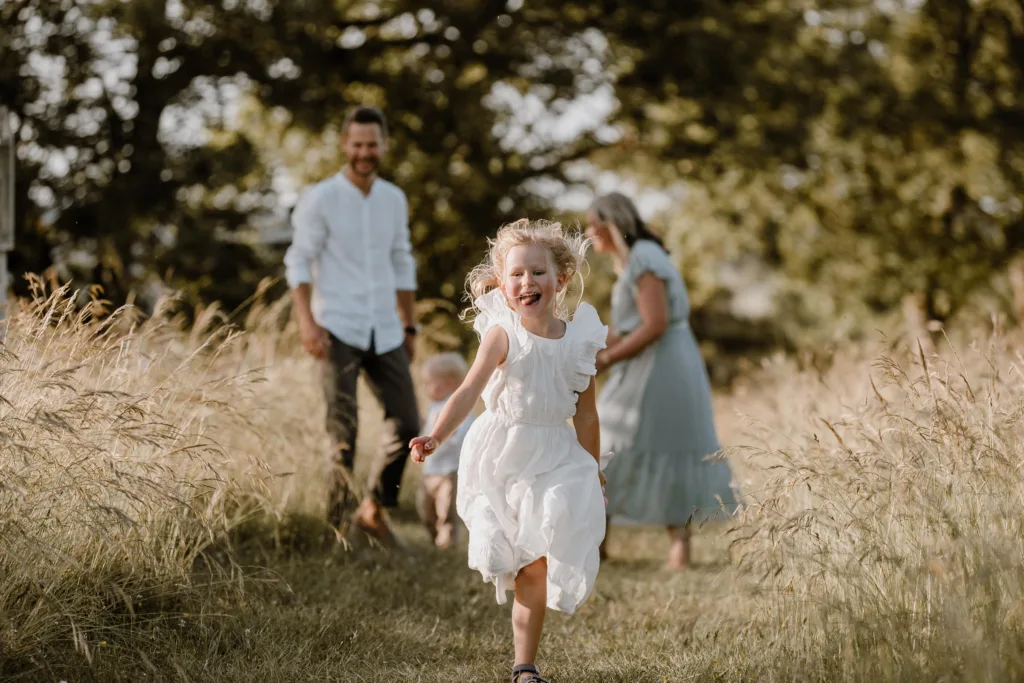 Lachendes Mädchen läuft durch eine Wiese, Eltern und Bruder folgen im Hintergrund