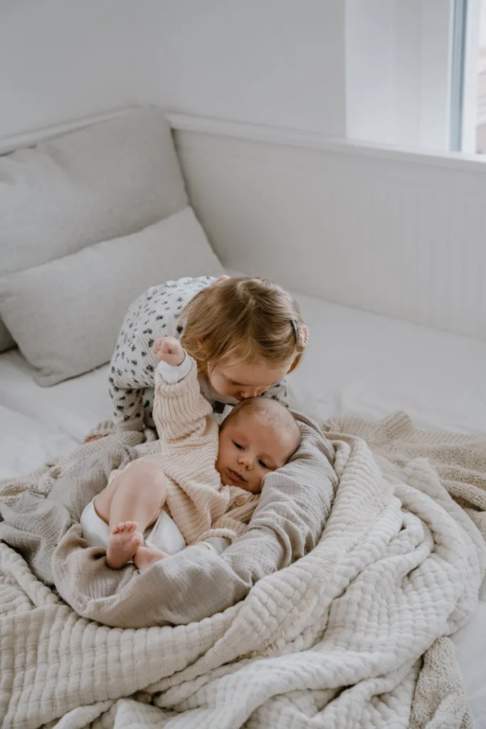 Kleines Mädchen gibt seinem Babygeschwisterchen auf einer weichen Decke auf dem Bett einen Kuss