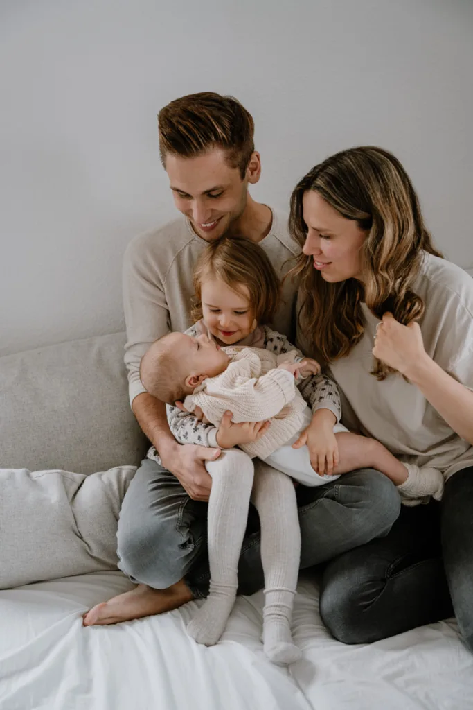 Junge Familie sitzt auf dem Bett. Die große Schwester darf das Baby halten