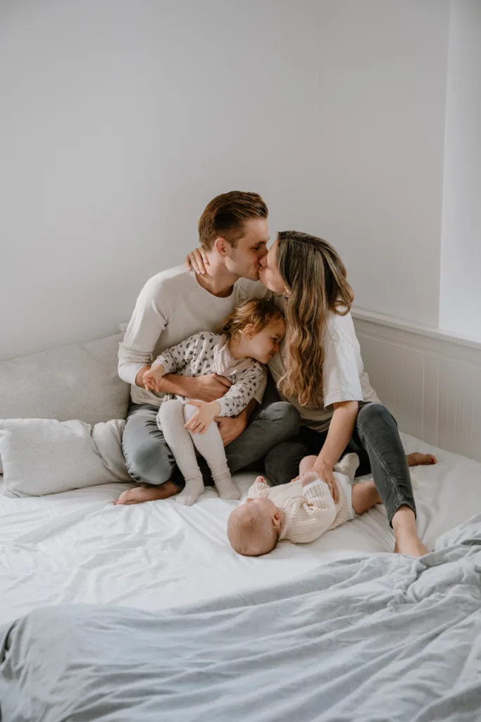 Junge Familie sitzt auf dem Bett. Die Eltern küssen sich. Das kleine Baby liegt vor ihnen und die große Schwester kuschelt sich an die Mutter.