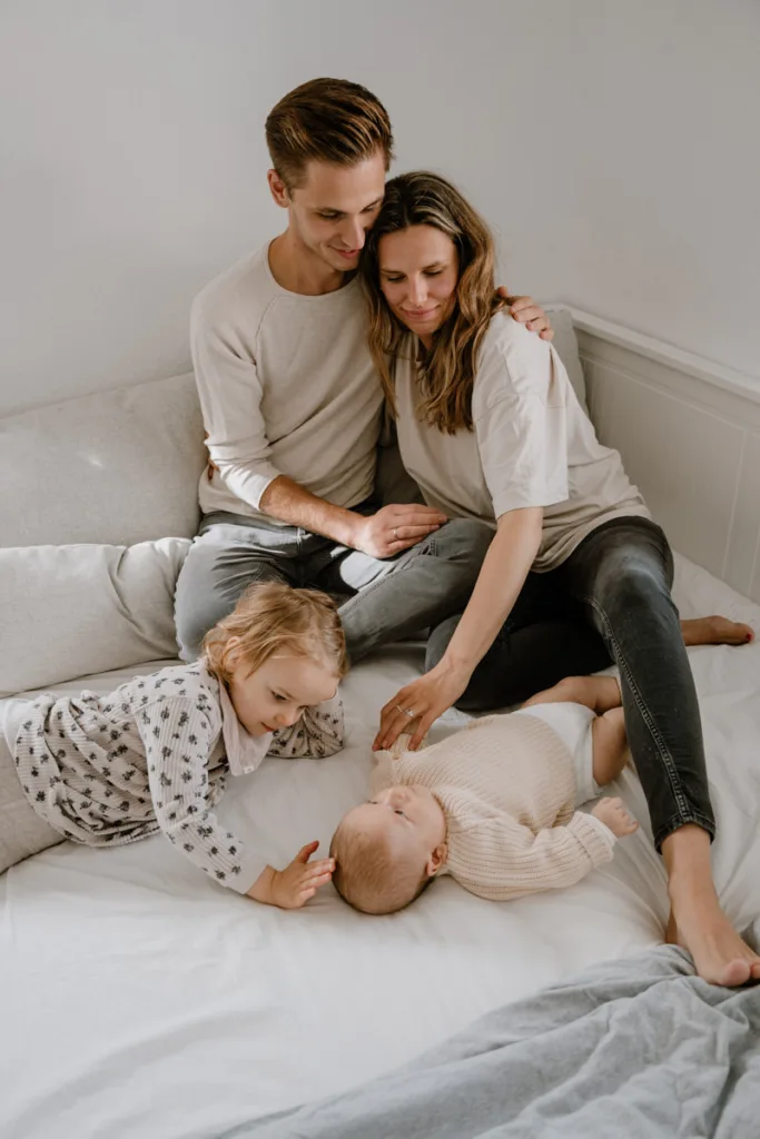 Familie sitzt auf dem Bett. Der Vater hält die Mutter im Arm und die große Schwester schaut über das Baby, welches vor ihr liegt an.