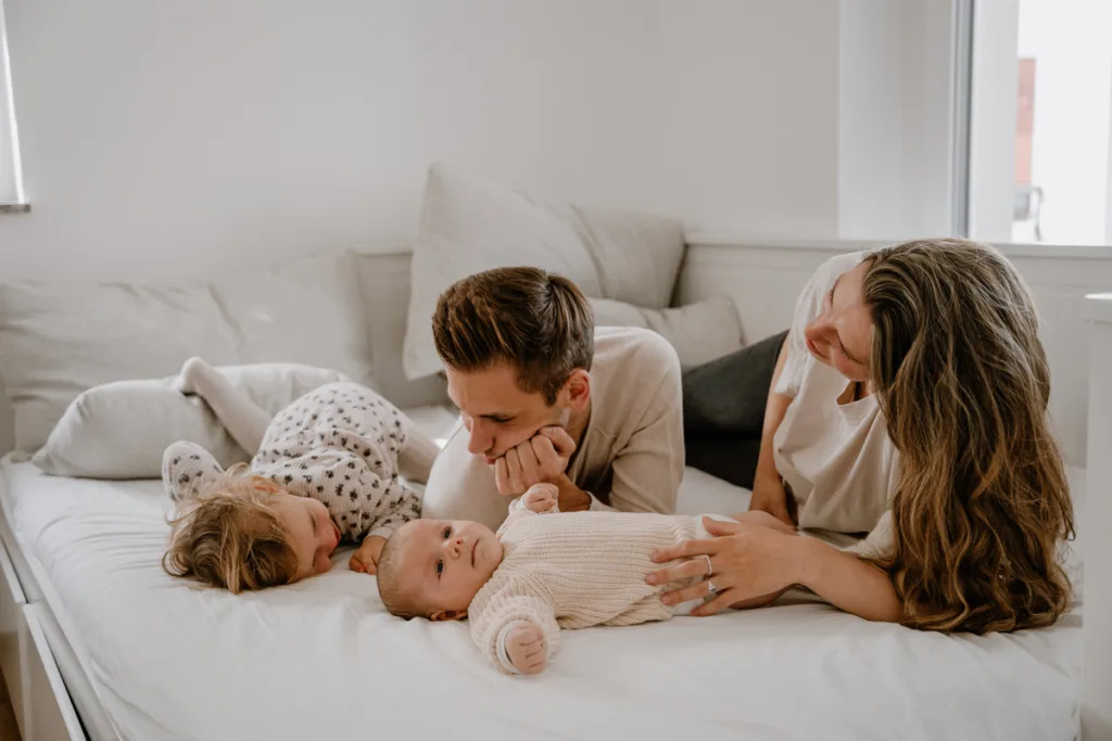 Junge 4- köpfige Familie liegt auf dem Bett. Das kleine Baby liegt vor ihnen und Mama und Papa berühren es mit ihren Händen.