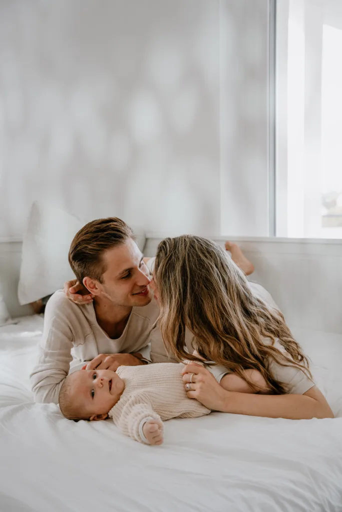 Eltern küssen sich im Bett während das Baby vor ihnen liegt und in die Kamera schaut.