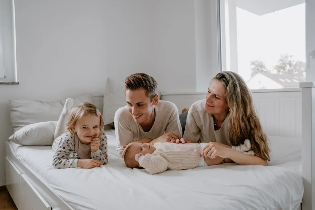 Junge 4- köpfige Familie liegt auf dem Bett. Das kleine Baby liegt vor ihnen und Mama und Papa berühren es mit ihren Händen.