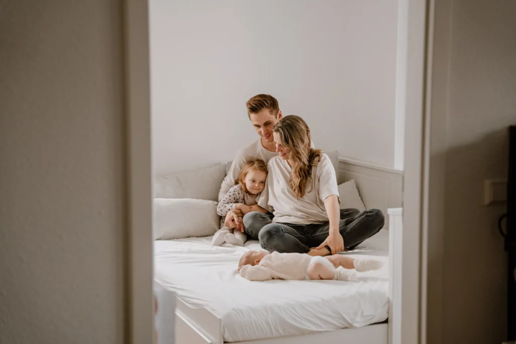 Junge 4- köpfige Familie sitzt auf dem Bett und schauen zum vor sich liegenden Baby hinab.