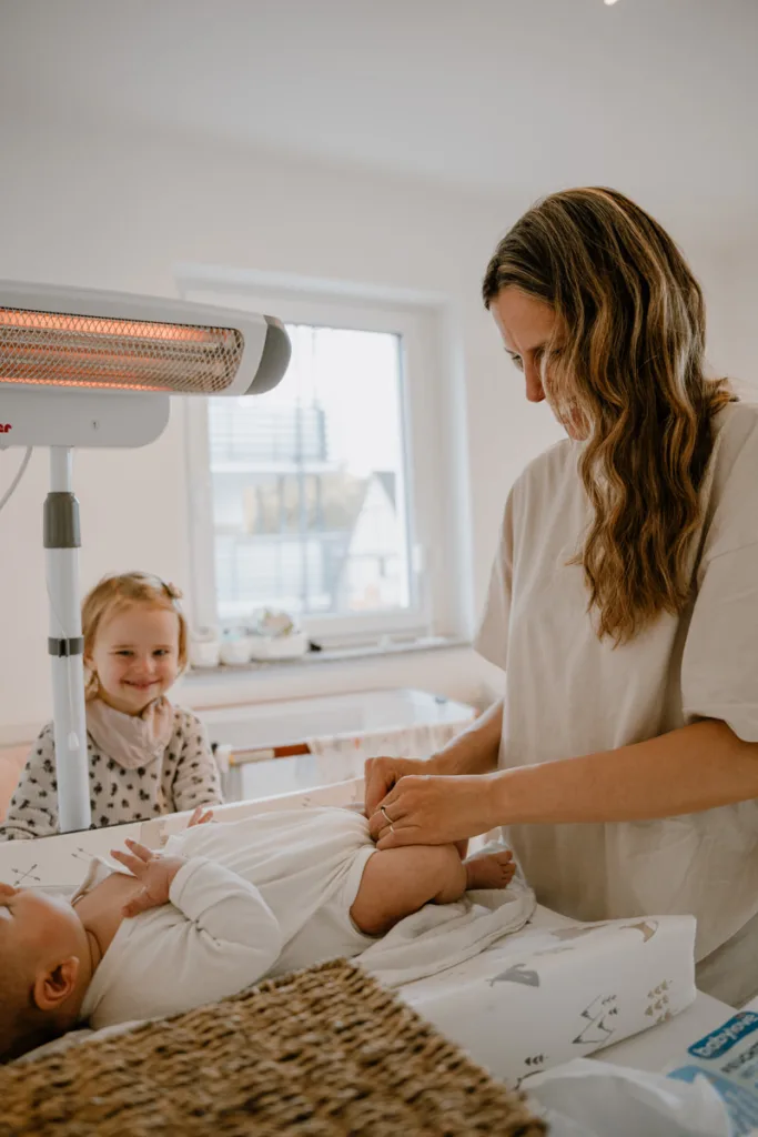 Mutter wickelt ihr Baby unter einer Wärmelampe, während die große Schwester daneben steht und lächelt