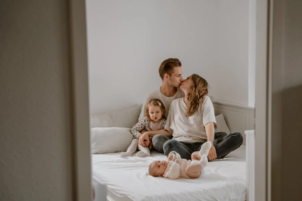 Junge Familie sitzt auf dem Bett. Die Eltern küssen sich und die große Schwester schaut auf das Baby, welches vor ihr auf dem Bett liegt.