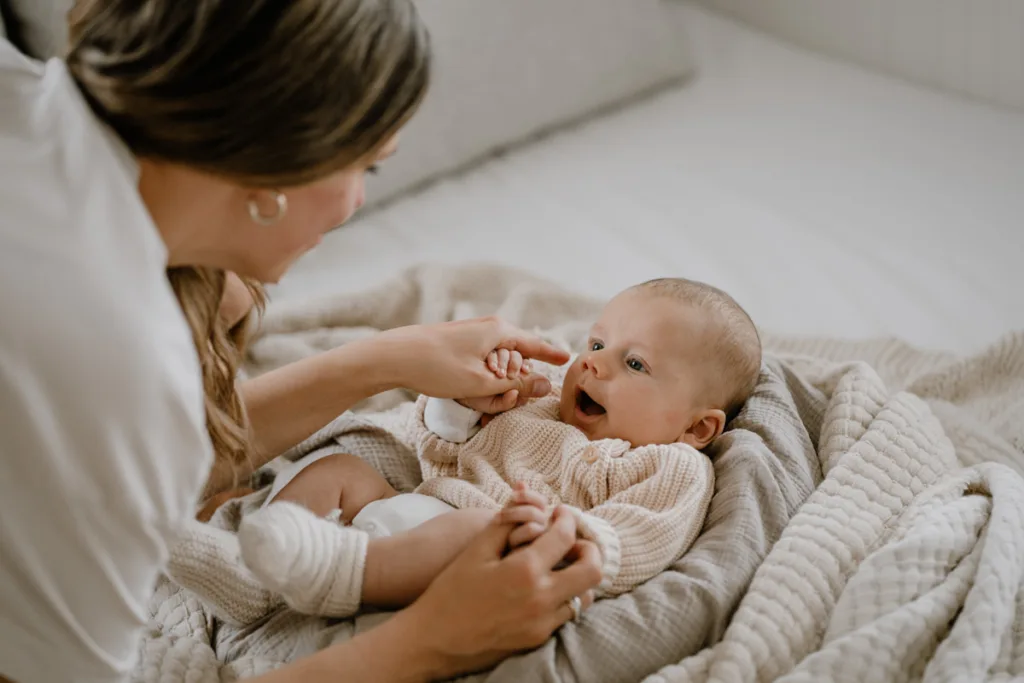 Ein Baby liegt lachend und entspannt auf weichen Decken und schaut mit großen Augen zu seiner Mama, die sich über ihn beugt.