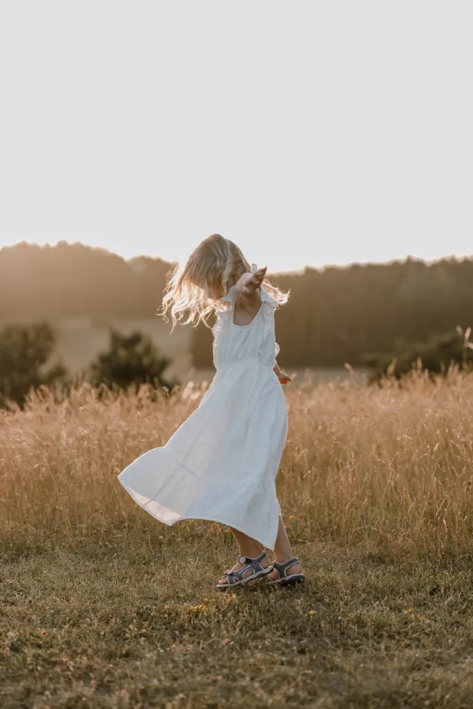 Kleines Mädchen im weißen Kleid dreht sich spielerisch auf einer Wiese im warmen Abendlicht
