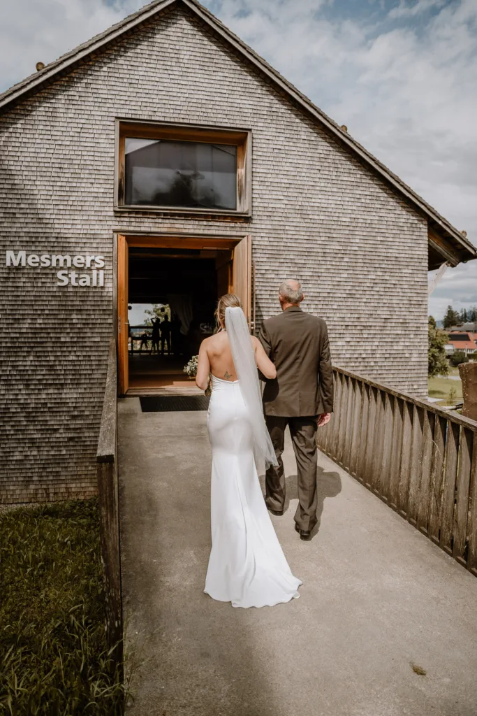 Braut wird von einem Mann in die Hochzeitslocation Mesmers Stall geführt, Blick von hinten.