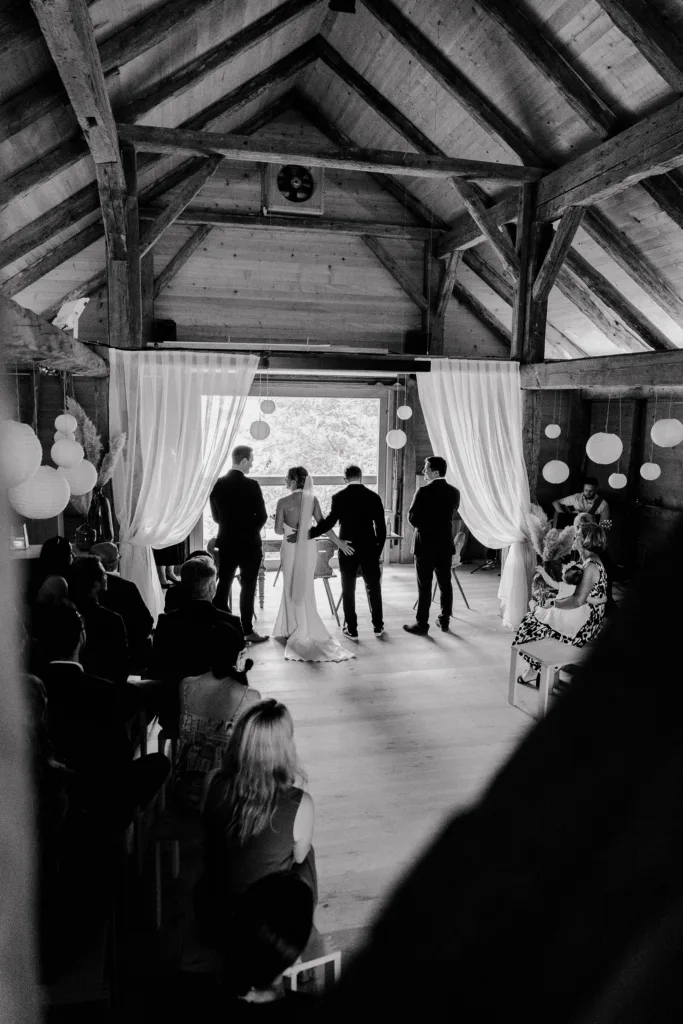 Brautpaar steht gemeinsam vorne bei der Trauung, aufgenommen von hinten in Schwarzweiß.