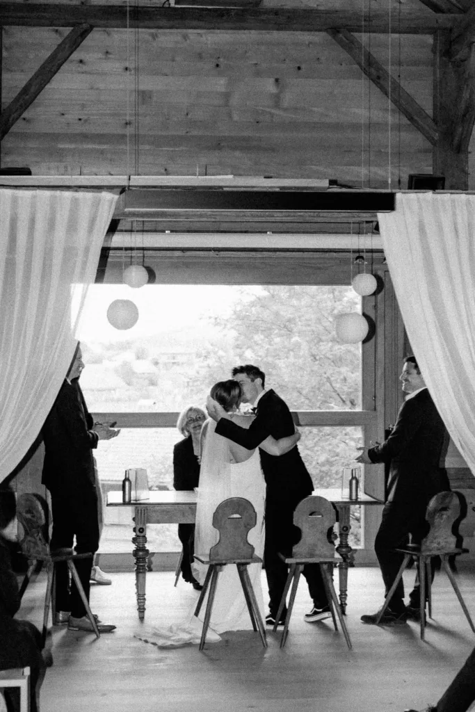 Schwarzweißaufnahme des Brautpaares beim Kuss während der Trauung, Gäste stehen ringsum.