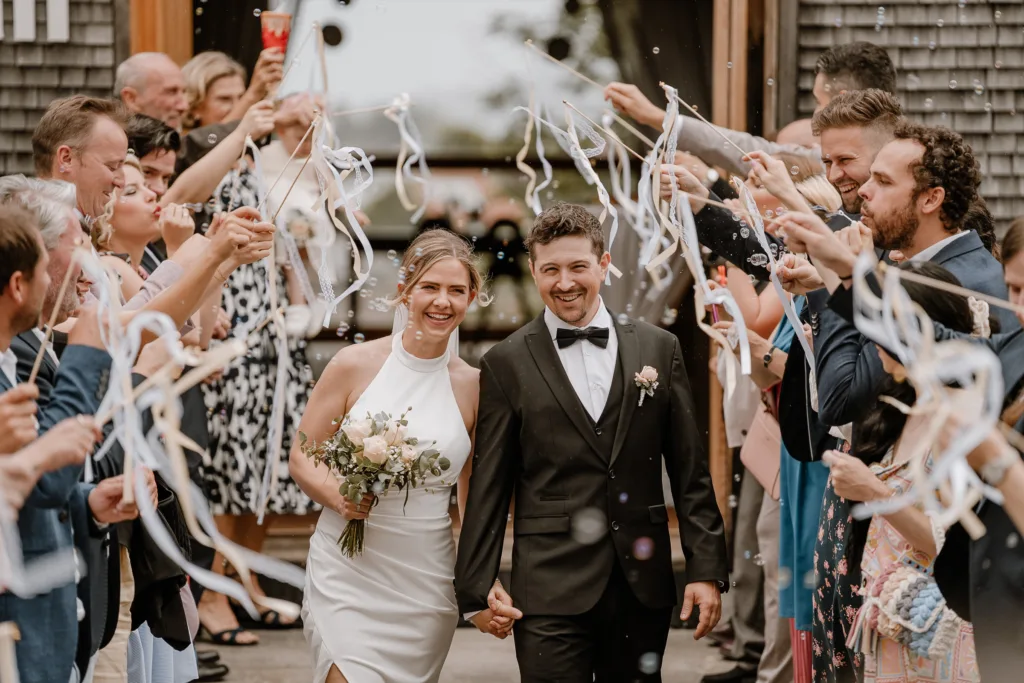 Brautpaar verlässt die Hochzeitslocation, Gäste bilden ein Spalier mit Bändern und Seifenblasen.