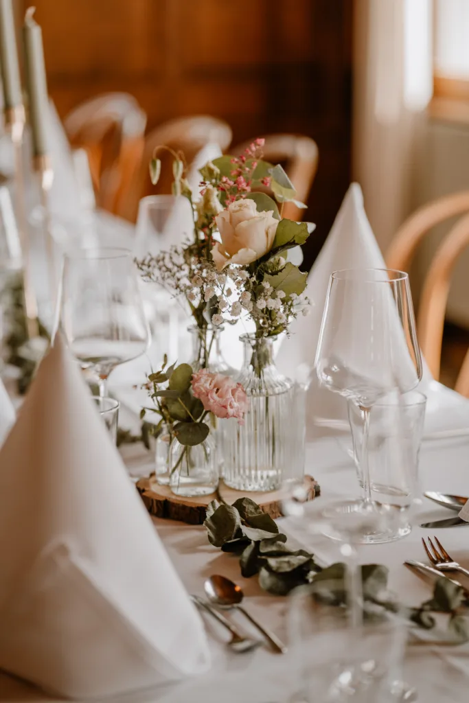 Hochzeitstafel mit Blumenvasen, Gläsern und Besteck in einem rustikalen Saal.