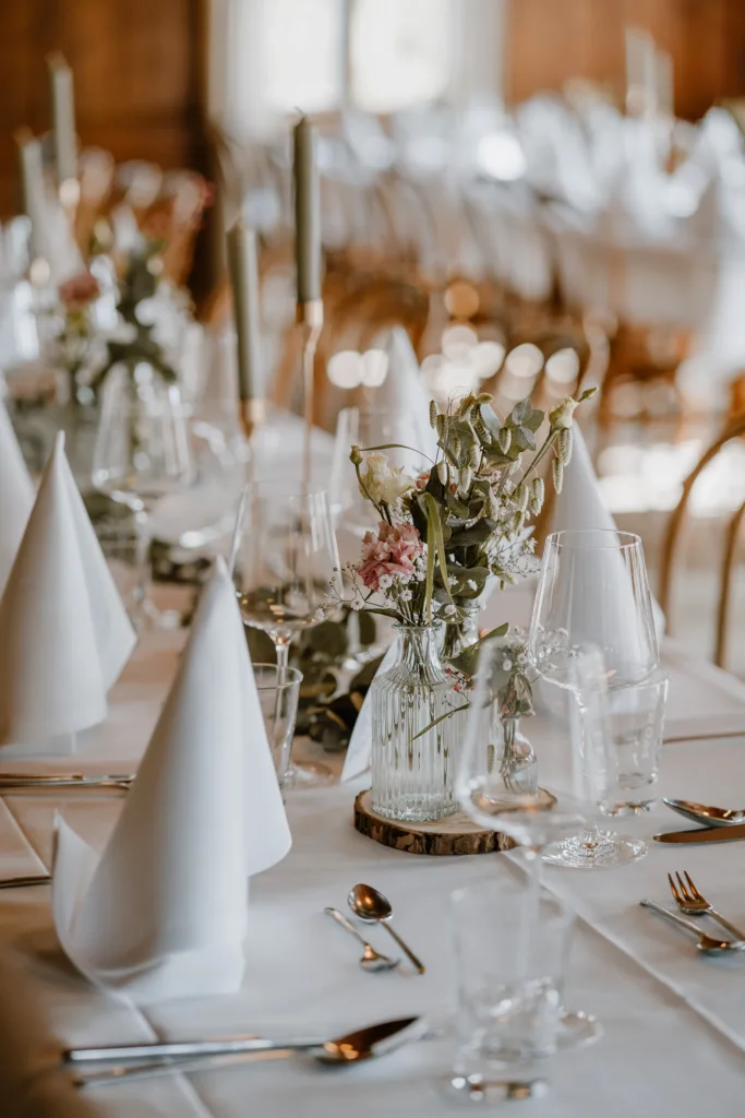 Hochzeitstafel mit Blumenvasen, Gläsern und Besteck in einem rustikalen Saal.