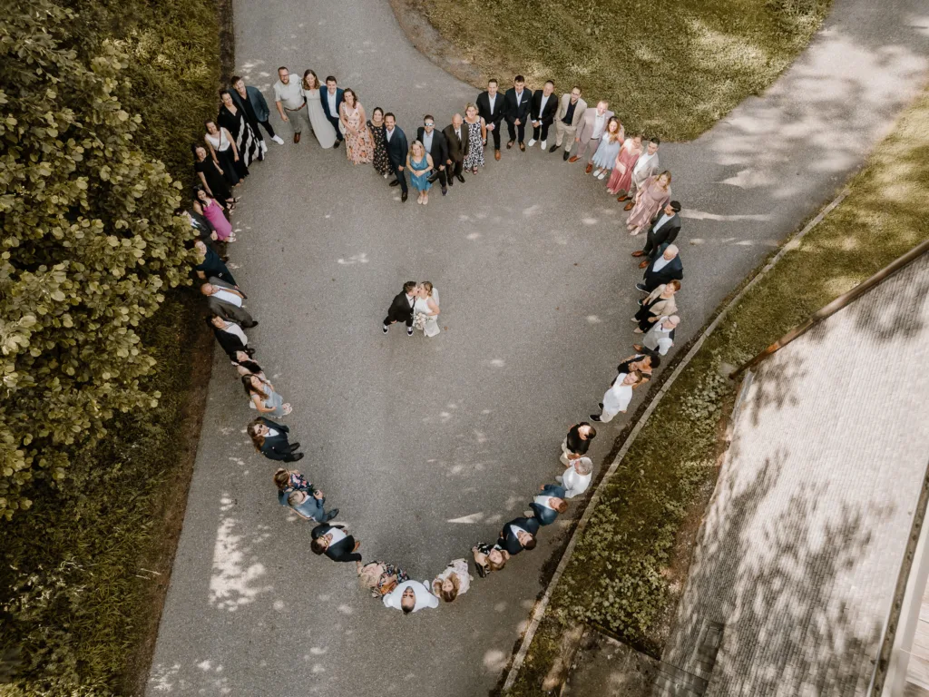 Drohnenaufnahme von Gästen, die ein Herz um das Brautpaar bilden.