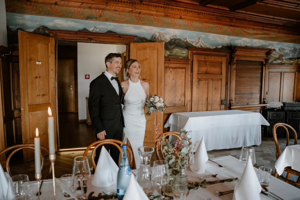 Brautpaar steht lachend im Hochzeitsaal neben der festlich gedeckten Tafel.