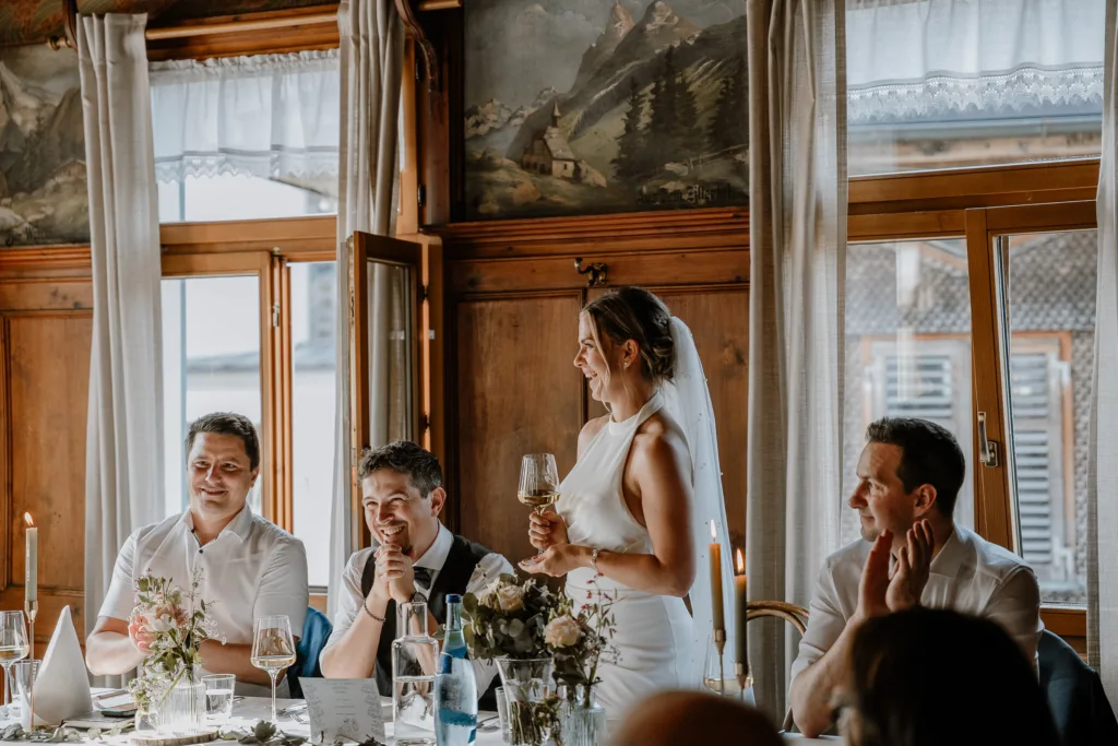 Braut hält eine Rede am Hochzeitstisch, Gäste hören aufmerksam zu.