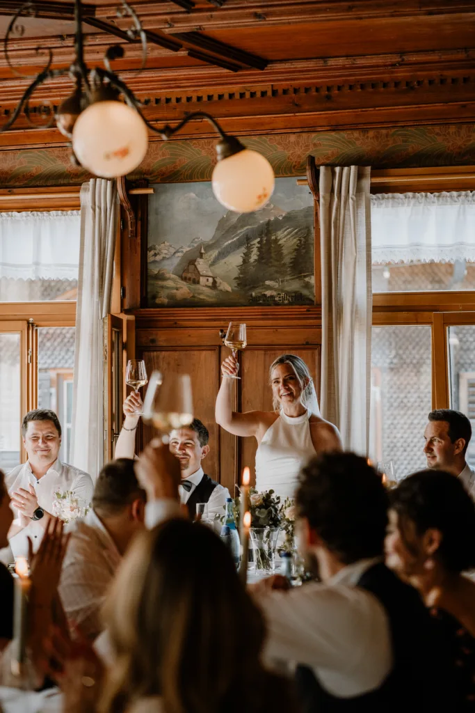 Braut erhebt das Weinglas zum Toast, Gäste stoßen gemeinsam an.