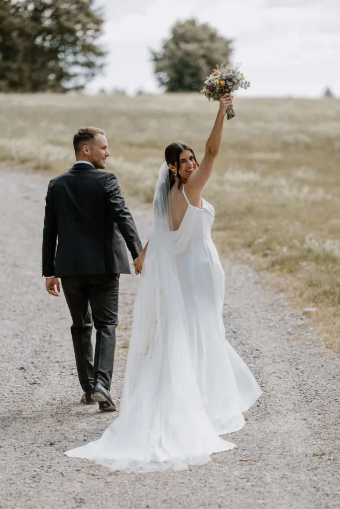 Braut hebt Brautstrauß jubelnd in die Luft auf Feldweg