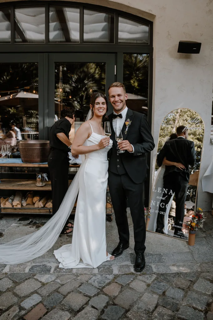 Brautpaar steht mit Champagnergläsern vor der Hochzeitslocation