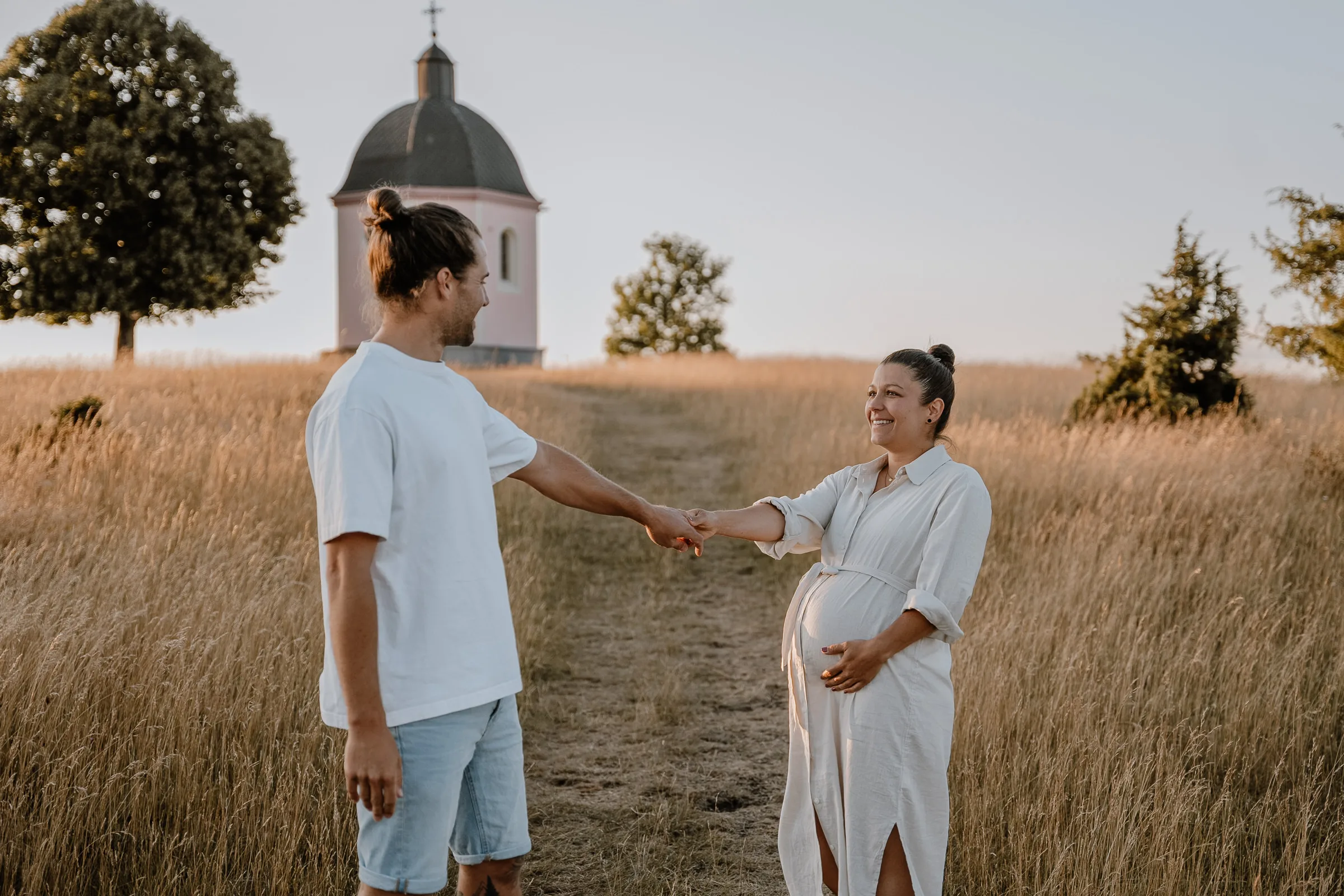 Paar bei der Babybauchfotografie hält sich an den Händen auf einem Feldweg, die werdende Mutter mit sichtbarem Babybauch